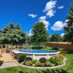 An above ground pool with a wooden deck attached to one side in a green backyard.