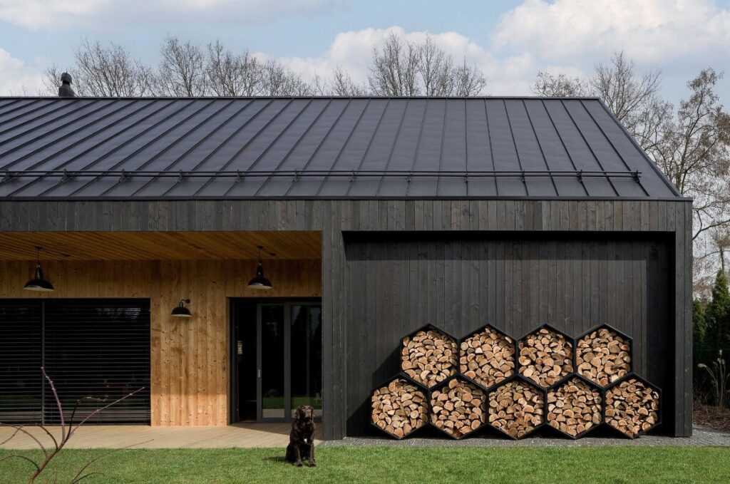 A modern black pole barn with dark metal roofing and a stack of firewood.