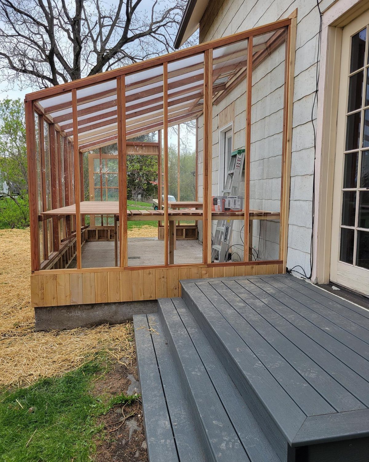 A wooden lean-to greenhouse frame under construction against a white house wall next to a deck.