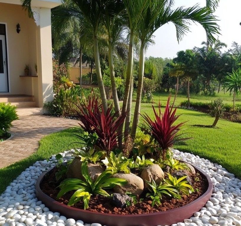 A circular garden bed with palm trees, red plants, and white pebbles on a green lawn.